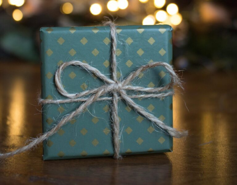 A decorated Christmas gift with a bow in front of warm bokeh lights and a tree.